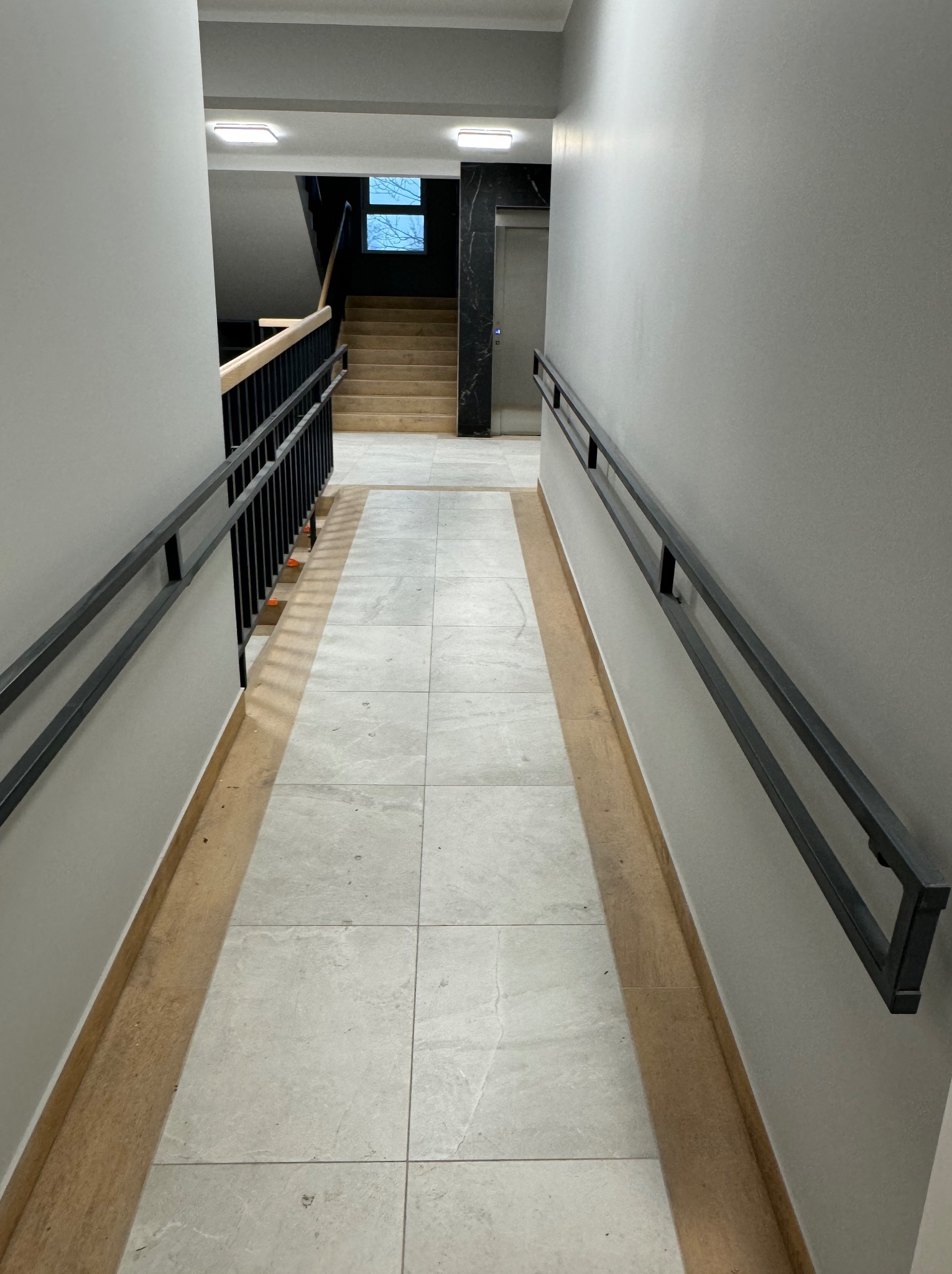Wnętrze korytarza z szarymi ścianami, jasnymi płytkami podłogowymi i czarnymi poręczami, prowadzącego do schodów i windy w budynku.