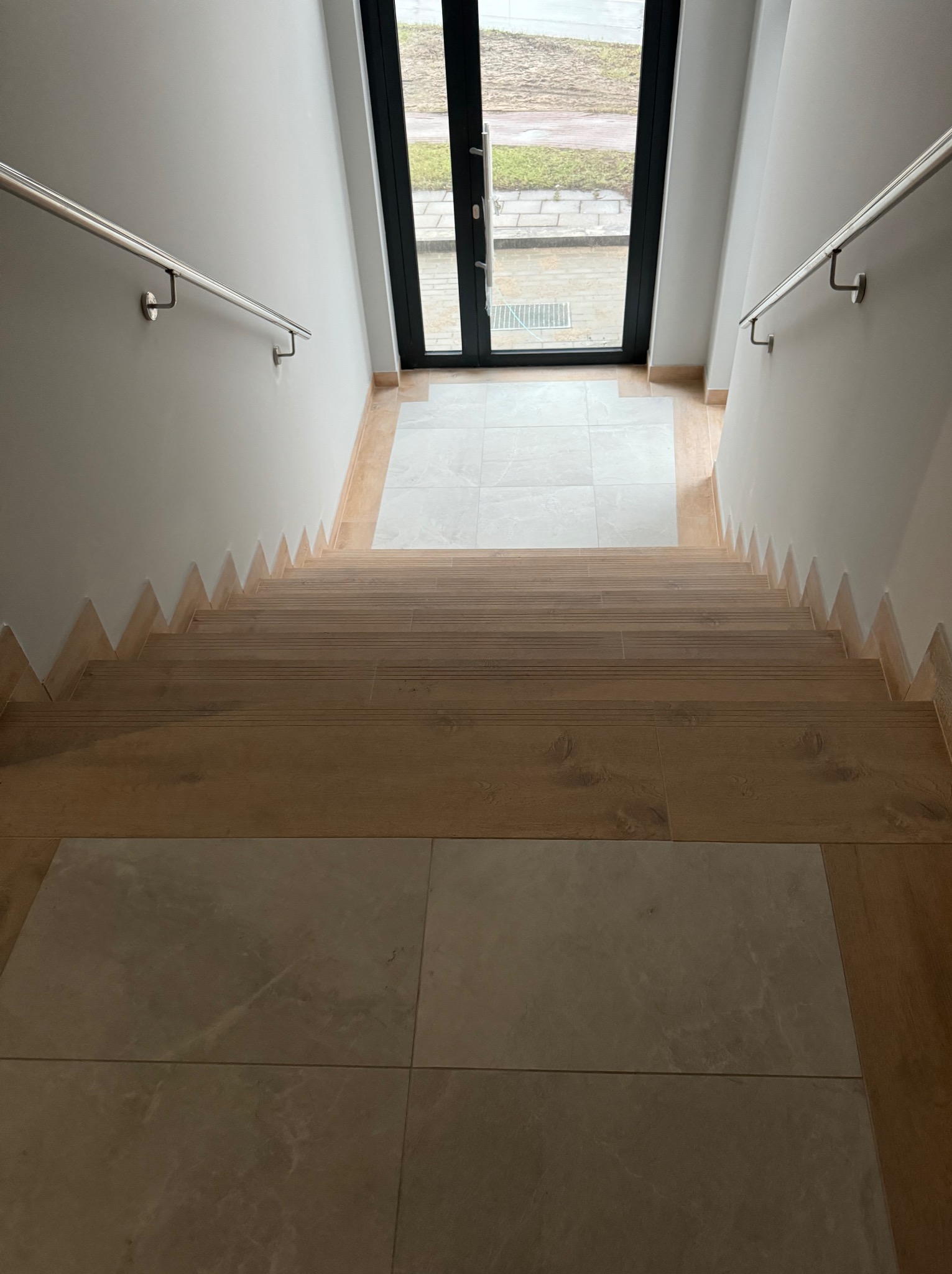 Schody wewnętrzne wykończone drewnopodobnymi panelami, z metalową balustradą i płytkami ceramicznymi u dołu oraz przy drzwiach wejściowych z przeszkleniami.