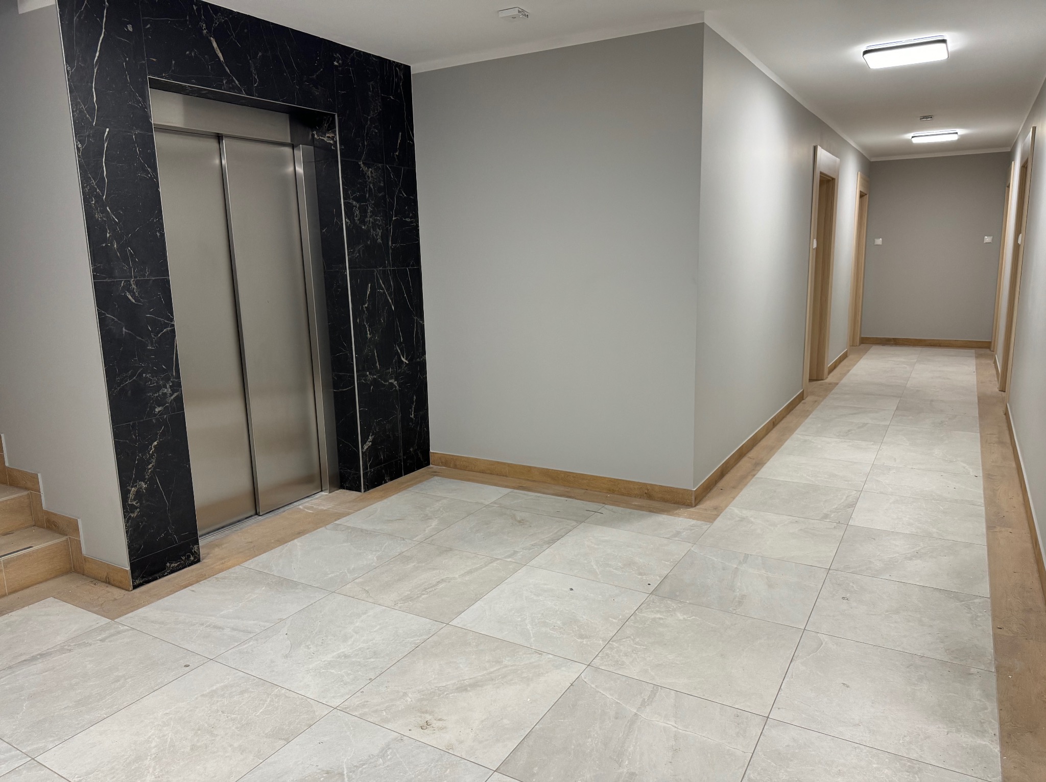 Korytarz w budynku z wykończonymi ścianami w szarym kolorze, jasnymi płytkami podłogowymi, wykończeniem przypodłogowym w kolorze drewna i windą obudowaną czarnym marmurem.