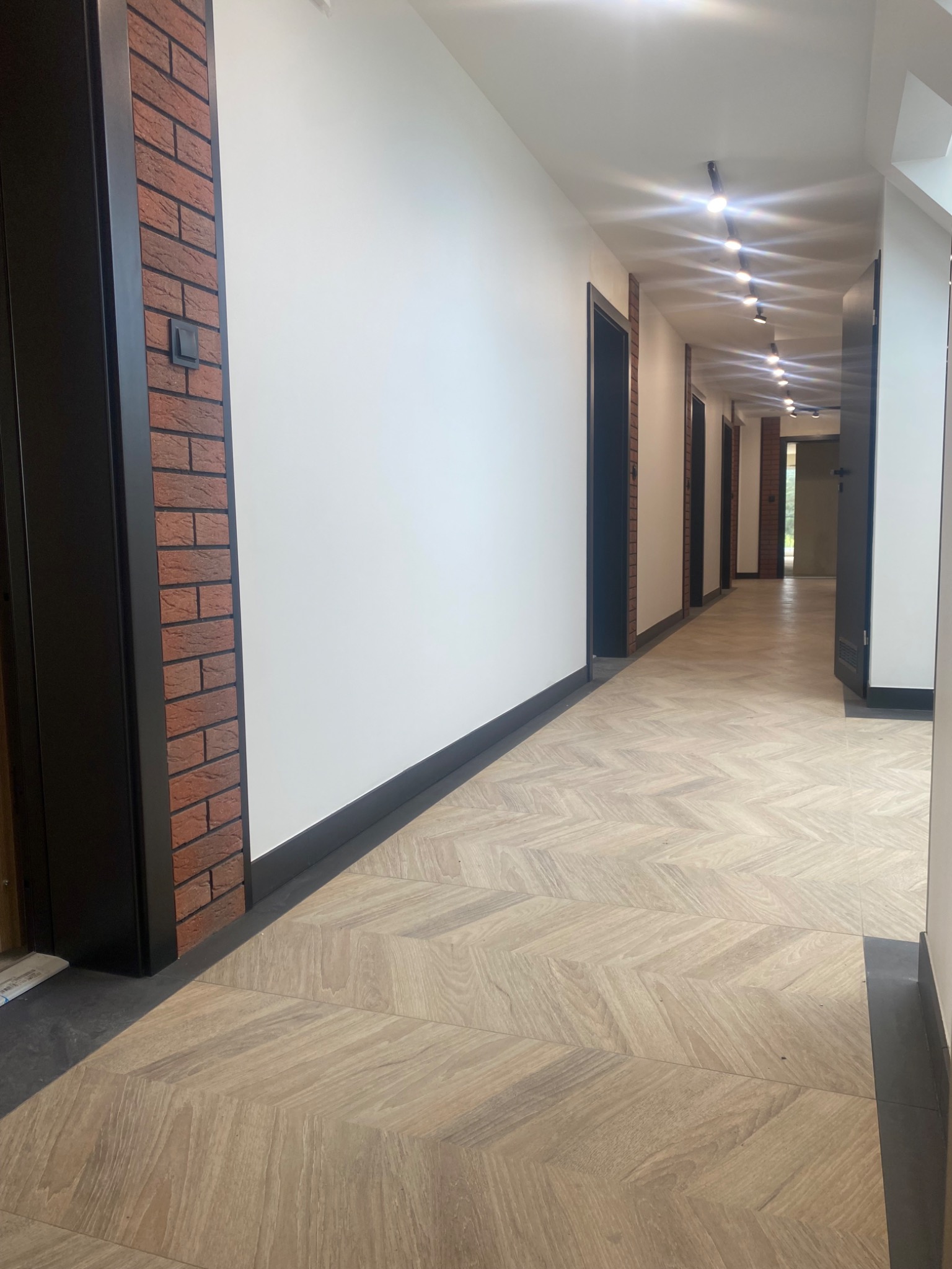 Długi korytarz z podłogą ułożoną w jodełkę, ściany białe, oświetlenie punktowe, drzwi z czarnymi futrynami i fragment dekoracyjnej ściany wyłożonej cegłą.