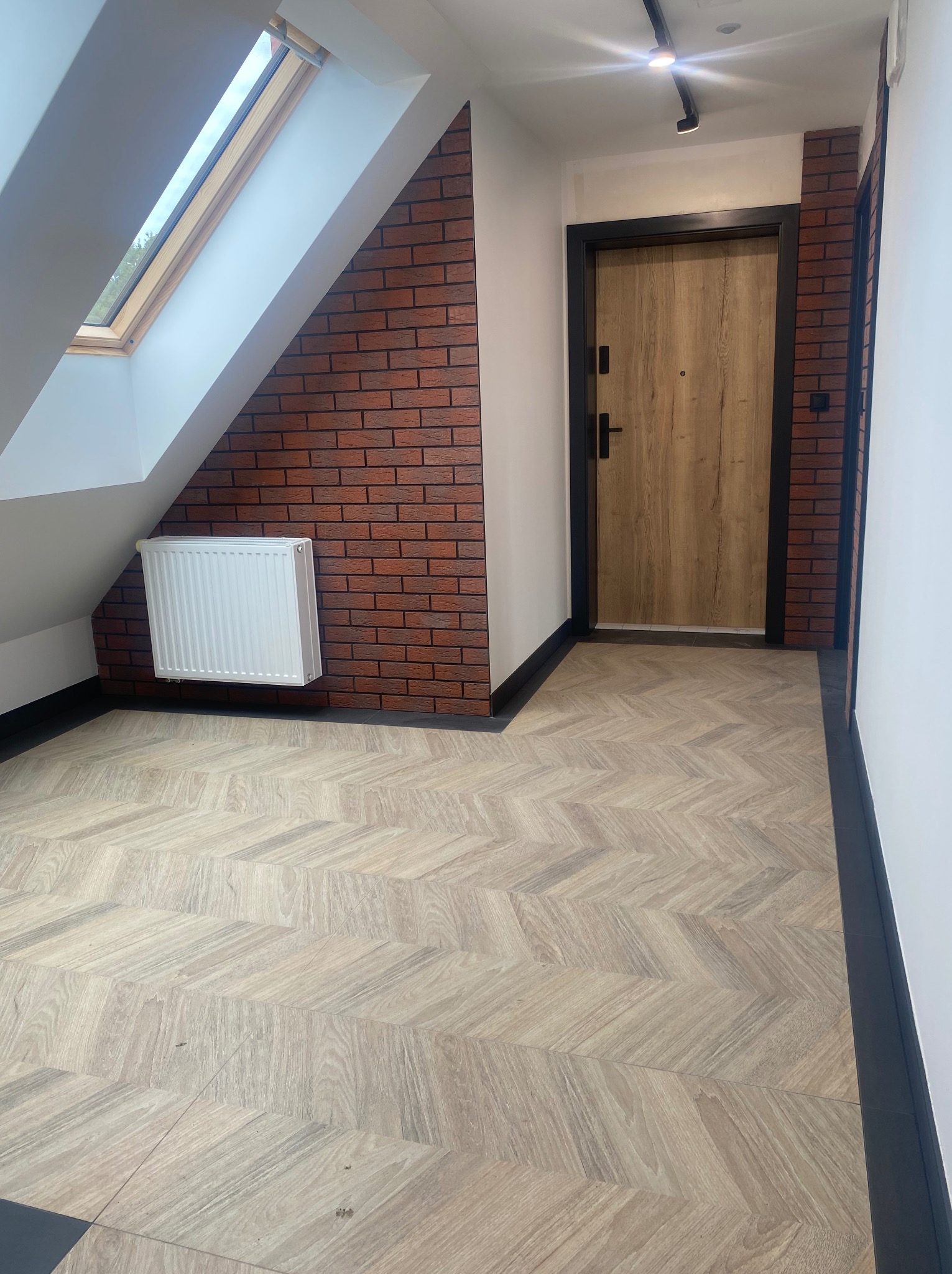 Wykończony przedpokój na poddaszu z drewnianą podłogą ułożoną w jodełkę, ceglaną ścianą, białym grzejnikiem i drewnianymi drzwiami z czarną ościeżnicą.