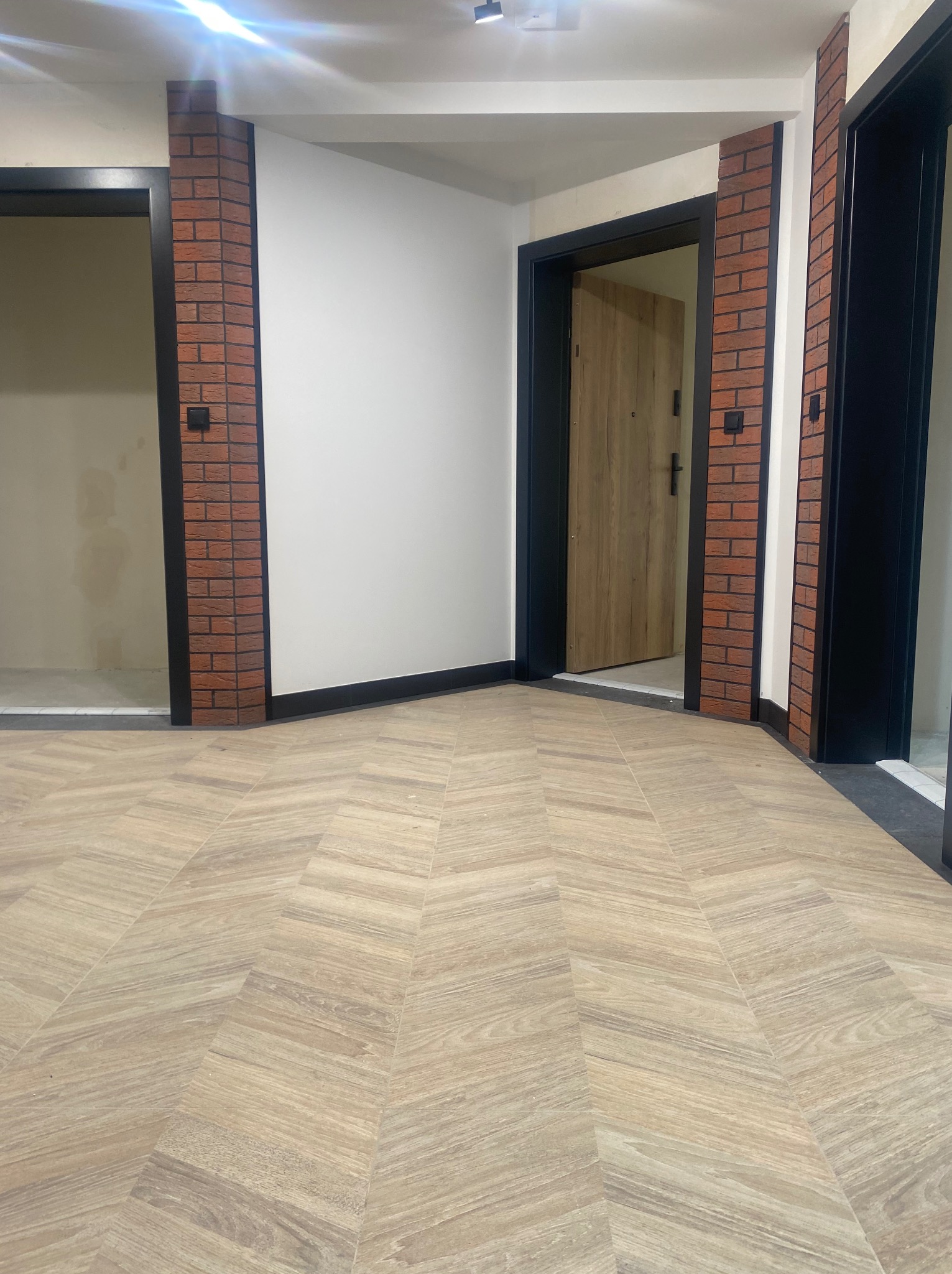 Wnętrze korytarza z trzema otworami drzwiowymi, wykończone panelami podłogowymi w jodełkę, obramowania drzwi w kolorze czarnym, z fragmentami cegieł dekoracyjnych po bokach.