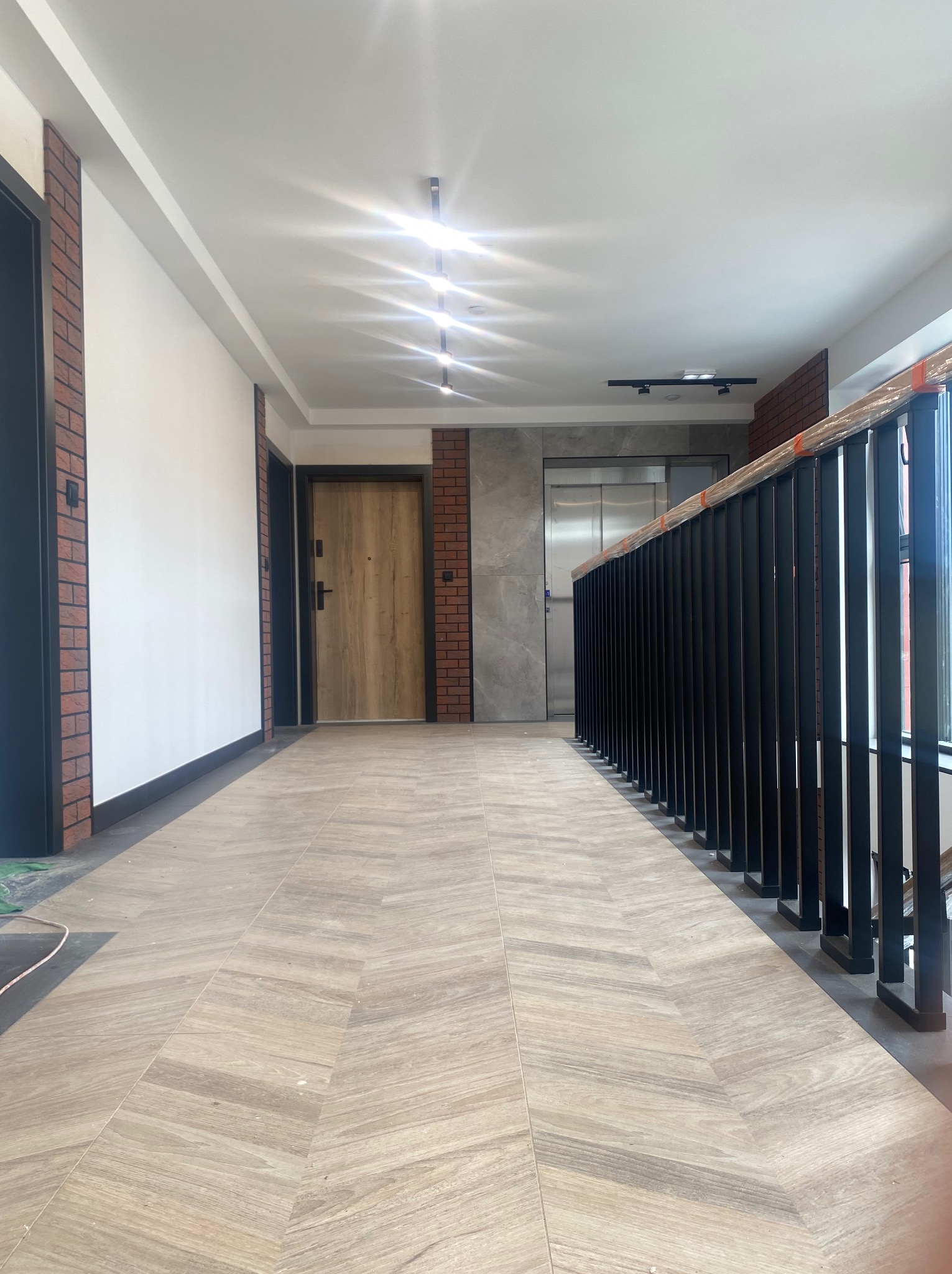 Korytarz w nowym budynku z wykończoną podłogą z paneli imitujących drewno ułożonych w jodełkę, czarne balustrady schodów zabezpieczone folią, drzwi wejściowe z drewnianą okleiną i fragment ściany...
