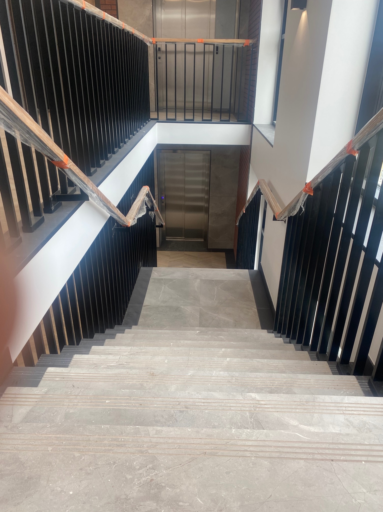 Wnętrze klatki schodowej w nowym budynku z marmurowymi schodami i czarnymi metalowymi balustradami, zabezpieczonymi folią ochronną na czas prac wykończeniowych; widoczne windy w tle.