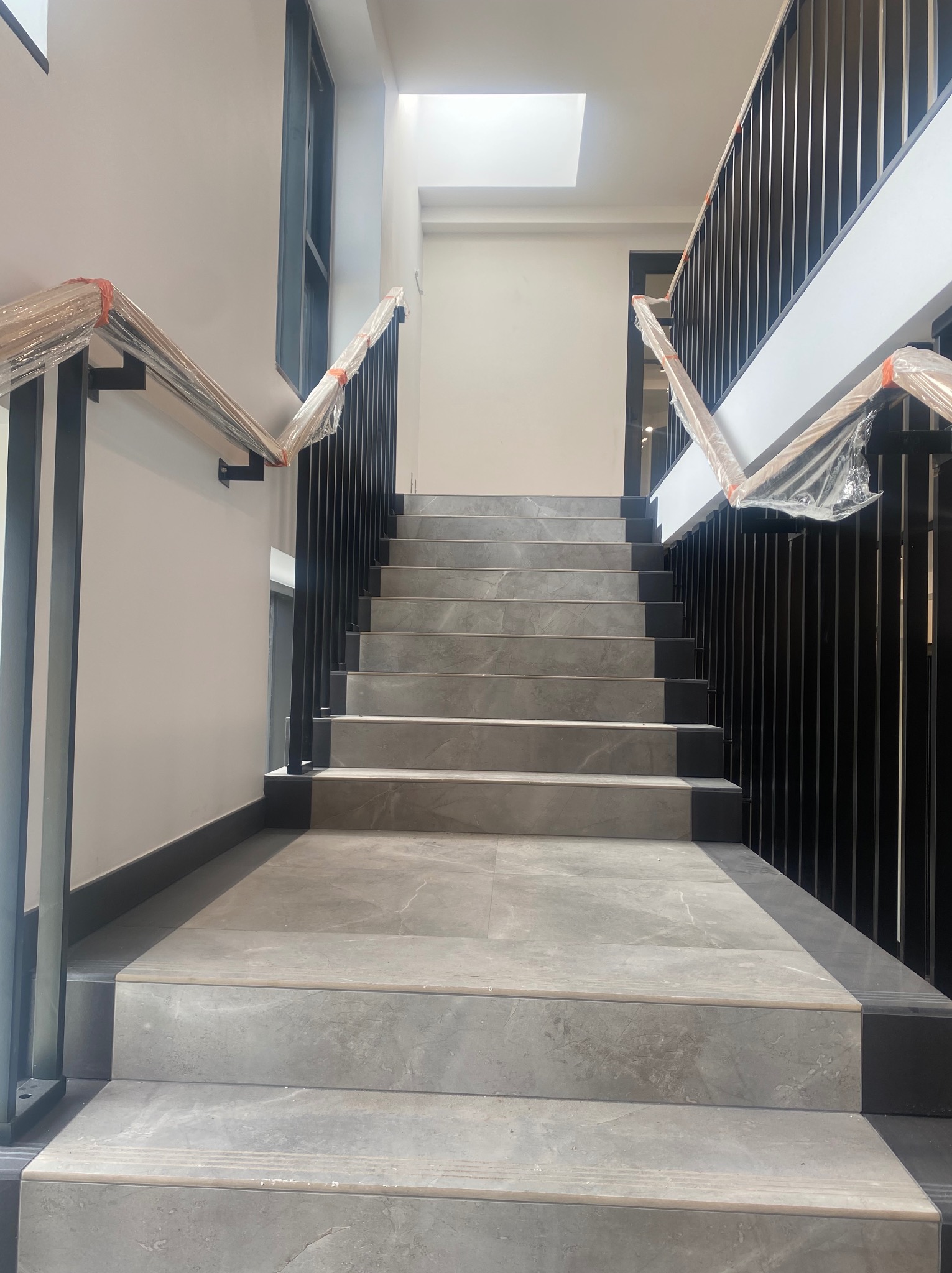 Schody wewnętrzne wyłożone szarymi płytkami imitującymi kamień, z czarną metalową balustradą, zabezpieczoną folią ochronną na poręczy.