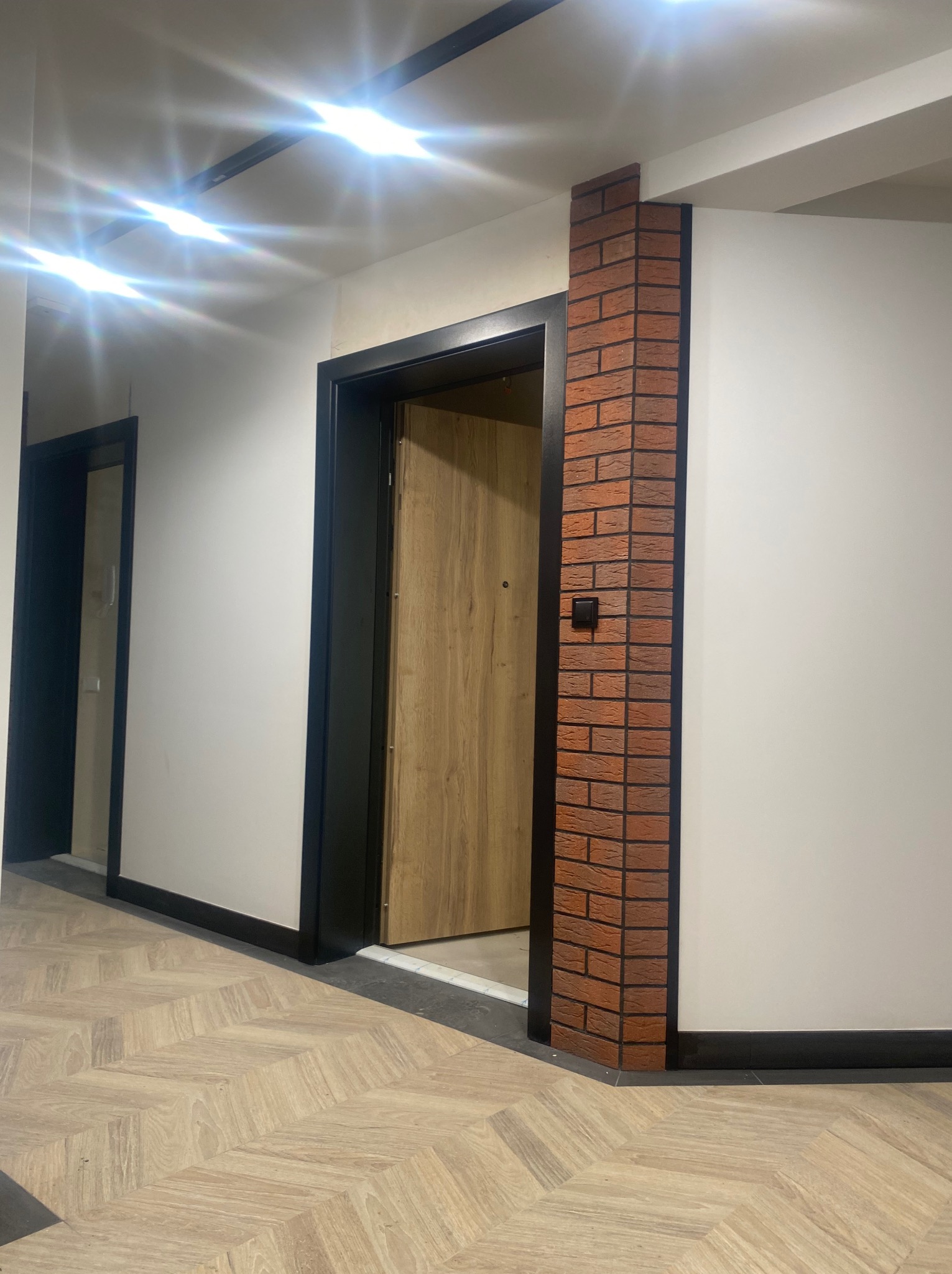 Korytarz z parkietem ułożonym w jodełkę, widoczne drzwi wejściowe z drewnianą okleiną i czarną ościeżnicą, fragment ściany wykończony cegłą klinkierową oraz nowoczesne oświetlenie sufitowe.