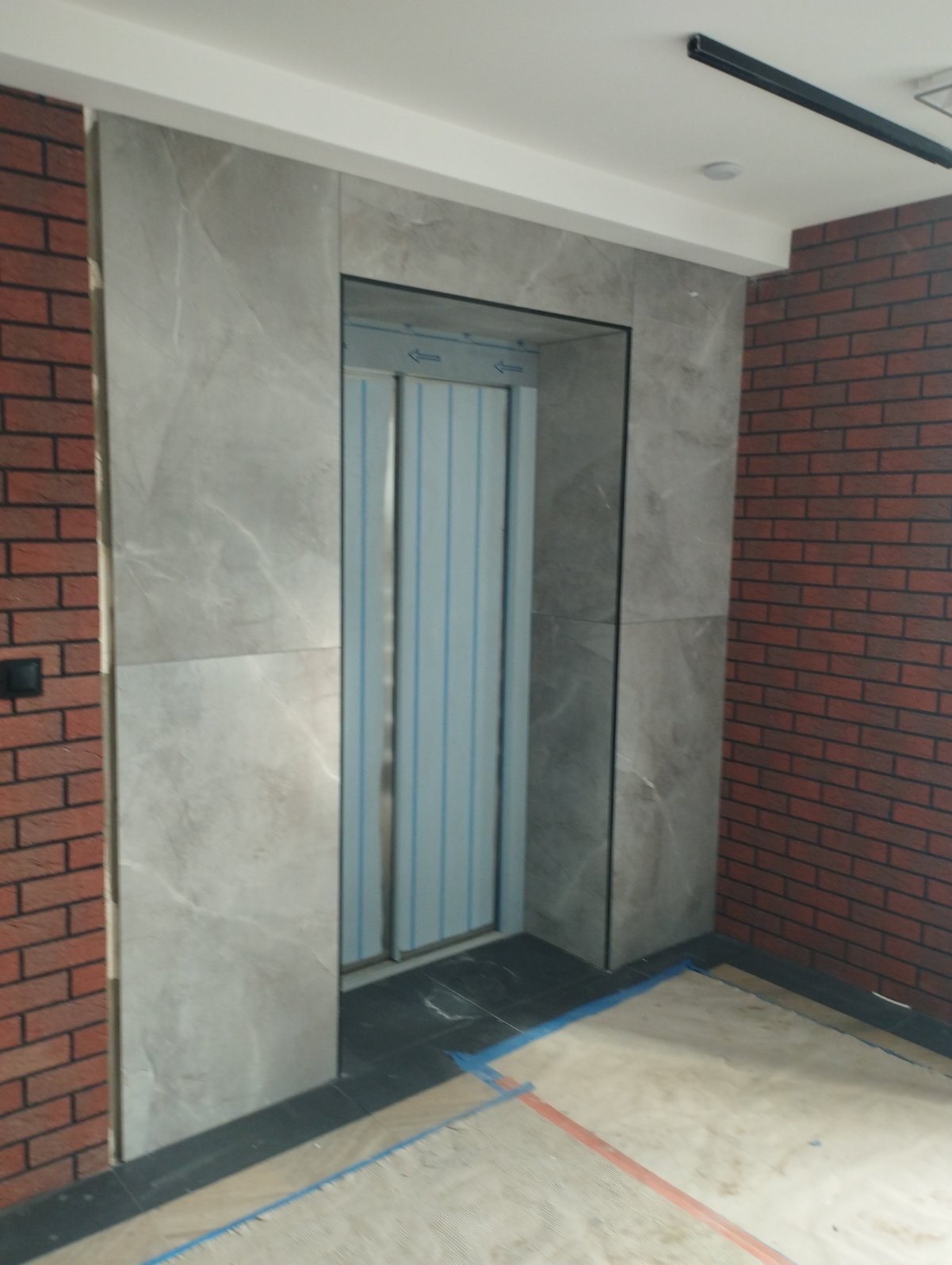 Wykończenie wnęki z windą w Świnoujściu: ściana z czerwonej cegły kontrastuje z marmurowymi płytami i metalowymi drzwiami windy.