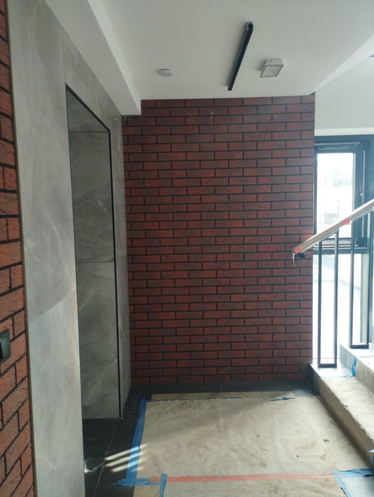Wnętrze budynku z fragmentami ścian wykończonymi czerwoną cegłą i szarym kamieniem, widoczne schody i zabezpieczona podłoga, oświetlenie sufitowe.