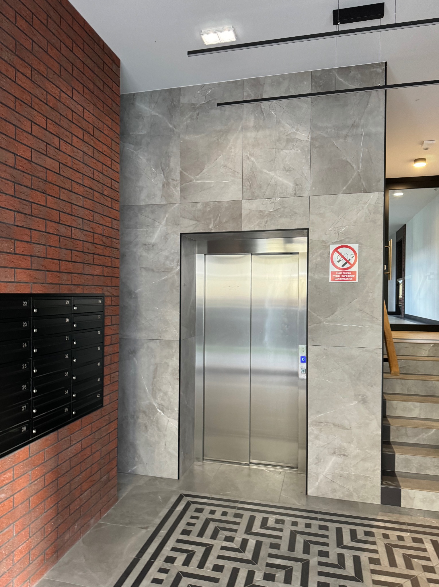 Nowoczesny hol wejściowy z windą, wykończony płytami imitującymi marmur i cegłą, z czarnymi skrzynkami pocztowymi i wzorzystą posadzką.