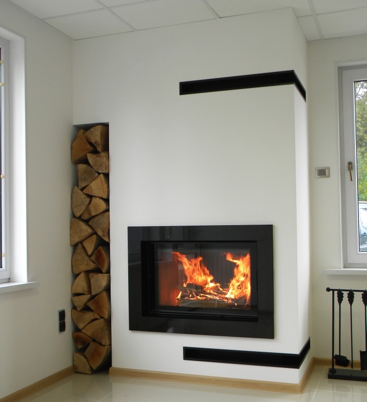 Nowoczesny kominek narożny z czarną obudową i płomieniami, obok wnęka z ułożonym drewnem, jasne ściany i podłoga.