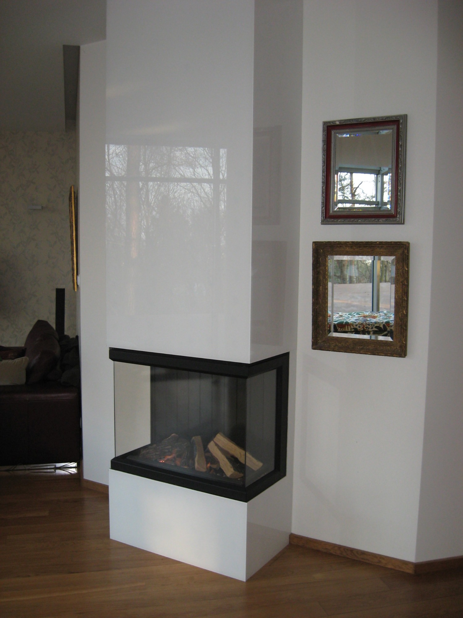 Nowoczesny narożny kominek obudowany błyszczącą białą płytą, z paleniskiem wypełnionym drewnem, widoczny w salonie z drewnianą podłogą, dwa lustra w ozdobnych ramach wiszą na ścianie obok.