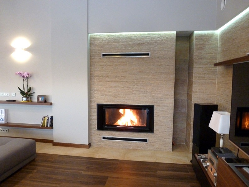 Nowoczesny salon z kominkiem w obudowie z beżowej cegły, płomienie widoczne przez szybę, dekoracyjne oświetlenie LED w suficie, drewniana podłoga i minimalistyczne półki z dekoracjami.