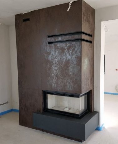 Nowoczesny kominek narożny z obudową imitującą rdzę, w surowym wnętrzu z widocznymi kablami i zabezpieczeniami podłogi.