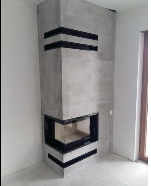 Surowy, narożny kominek z szarego betonu architektonicznego, z czarnymi wstawkami wentylacyjnymi, w trakcie montażu, widoczne ślady po wkrętach na powierzchni.