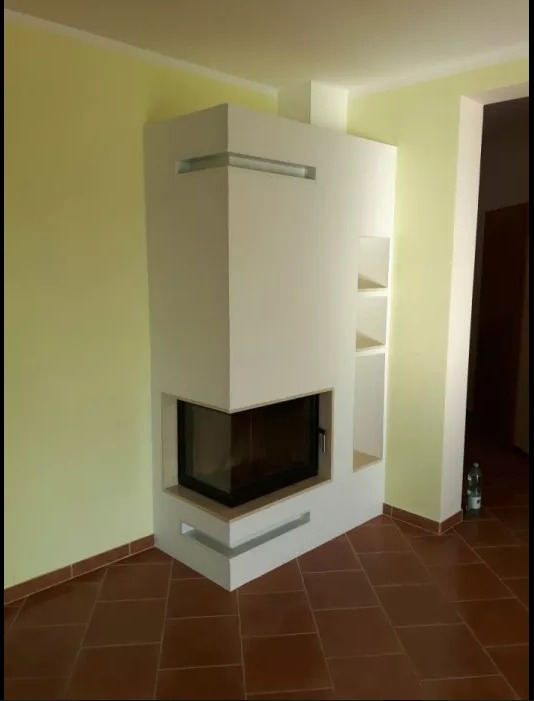 Nowoczesny, narożny kominek w obudowie z białego matowego MDF z półkami, na tle żółtej ściany i brązowej terakoty.