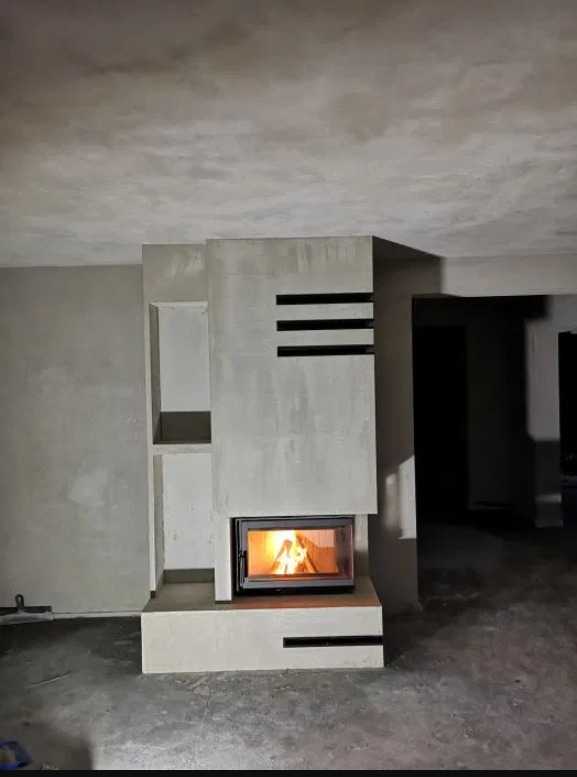 Surowy kominek z czarnymi akcentami w trakcie wykończenia, widoczne niedoskonałości tynku i surowy beton na podłodze, płomienie wewnątrz paleniska.