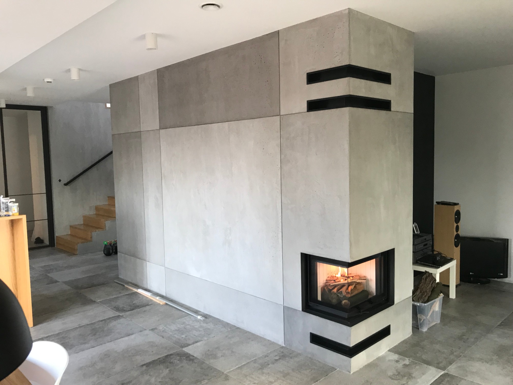Nowoczesny kominek narożny z szarego betonu architektonicznego z czarnymi akcentami, płomienie widoczne za szybą, na podłodze szare płytki imitujące kamień, w tle schody i sprzęt RTV.
