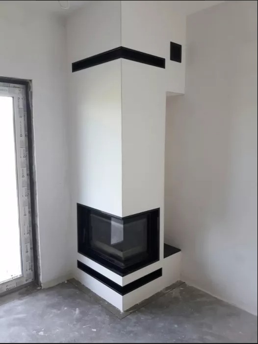 Biały narożny kominek z czarnymi elementami dekoracyjnymi w surowym wnętrzu, widoczna szyba paleniska i fragment okna z PCV.