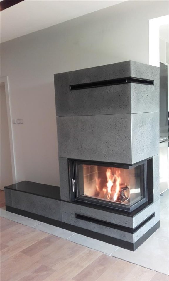 Nowoczesny kominek narożny z szarego betonu architektonicznego z czarnymi akcentami, płomienie widoczne za szybą, jasne drewniane panele podłogowe.