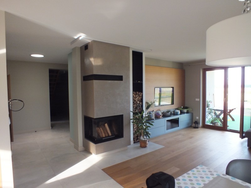 Nowoczesny salon z narożnym kominkiem obudowanym beżowym tynkiem strukturalnym, z czarnymi wstawkami i zapasem drewna obok. Widok na taras przez przesuwne drzwi.