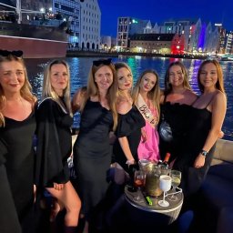 Yacht Club Premier - Grupa kobiet w czarnych sukienkach świętuje wieczór panieński na łodzi, w tle oświetlony port miejski i zabytkowy statek.
