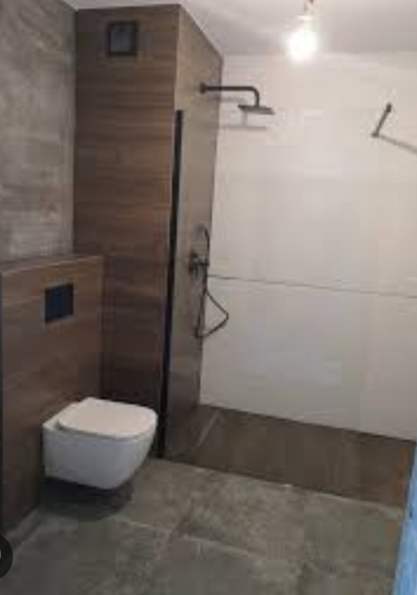 Nowoczesna łazienka z podwieszaną toaletą, ciemnymi płytkami imitującymi drewno na ścianie i białymi płytkami w strefie prysznica z czarną armaturą.