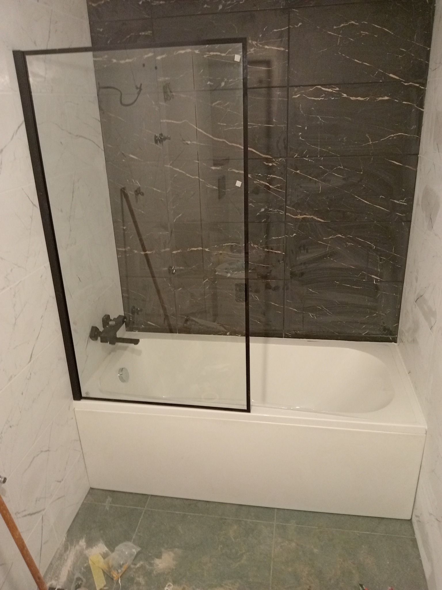 Nowoczesna łazienka z wanną obudowaną białą płytą, czarną armaturą i szklanym parawanem nawannowym z czarną ramą, płytki marmurowe na ścianach i podłodze, widoczne ślady po remoncie.