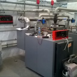 EKOSTAL - TECHNIKA GRZEWCZA KACZMAREK SEBASTIAN - Dwa szare piece grzewcze z żółtymi rurami gazowymi, zbiornik wyrównawczy w folii, widoczne elementy instalacji i rury na ścianie w kotłowni z białymi kafelkami.