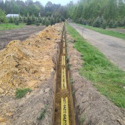 EKOSTAL - TECHNIKA GRZEWCZA KACZMAREK SEBASTIAN - Wykop z ułożoną żółtą rurą gazową w folii ochronnej, widoczne świeże ziemne skarpy i otoczenie terenu z młodymi drzewkami iglastymi.