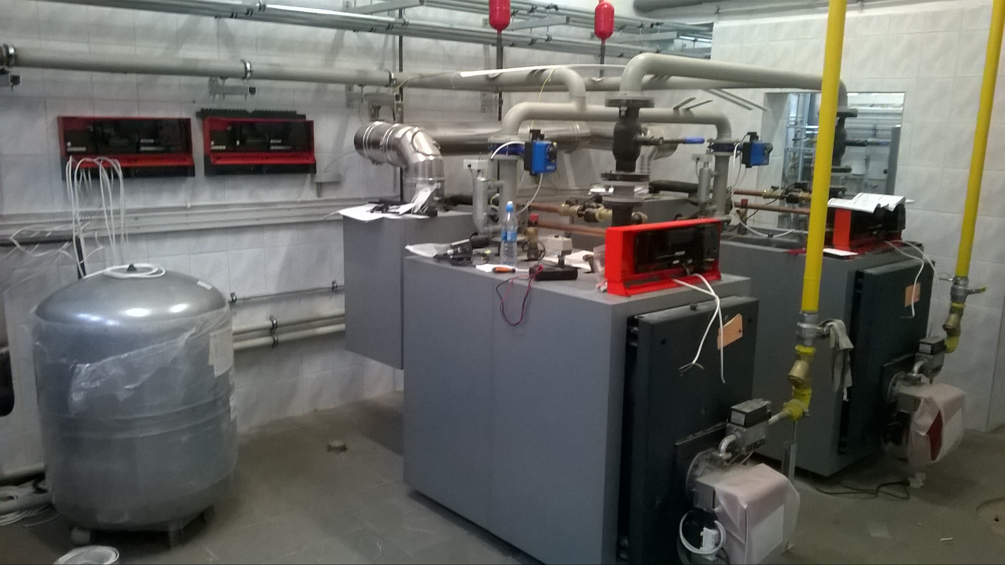 Dwa szare piece grzewcze z żółtymi rurami gazowymi, zbiornik wyrównawczy w folii, widoczne elementy instalacji i rury na ścianie w kotłowni z białymi kafelkami.