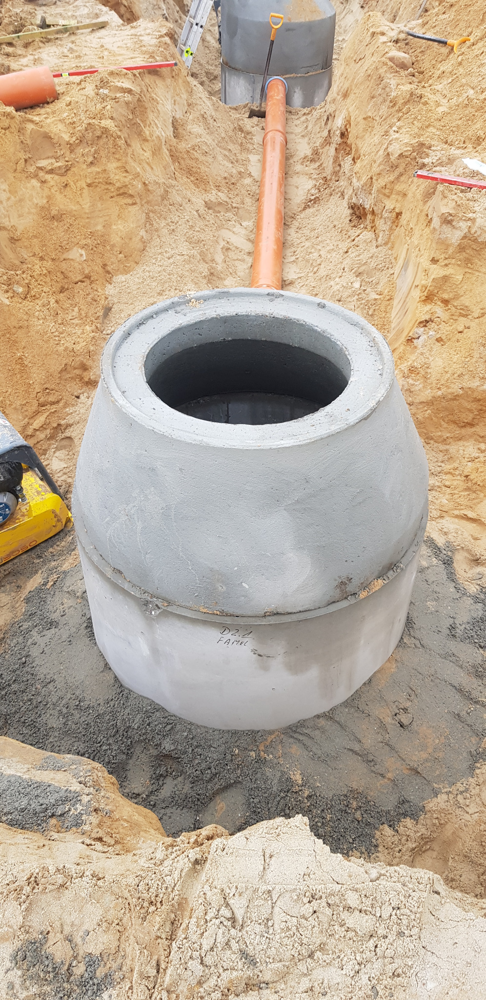 Betonowy zbiornik z włazem, prawdopodobnie szambo, umieszczony w wykopie z piasku, z widoczną pomarańczową rurą doprowadzającą ścieki.