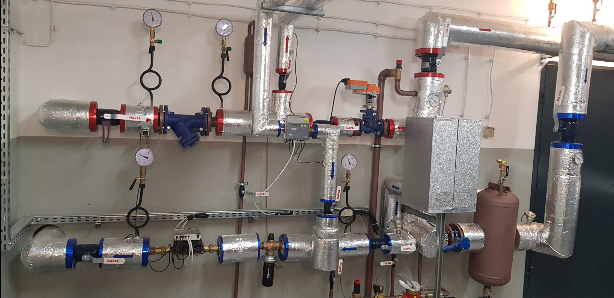 Złożona instalacja rur grzewczych z zaworami i manometrami, zaizolowana folią aluminiową, widoczne strzałki kierunku przepływu, filtry siatkowe oraz skrzynka sterownicza.