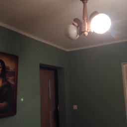 Mateusz Mazewski - Pokój z zielonymi ścianami, drewnianą lampą sufitową z dwoma kloszami i obrazem w ramie. Widoczny fragment drzwi i sufitu.