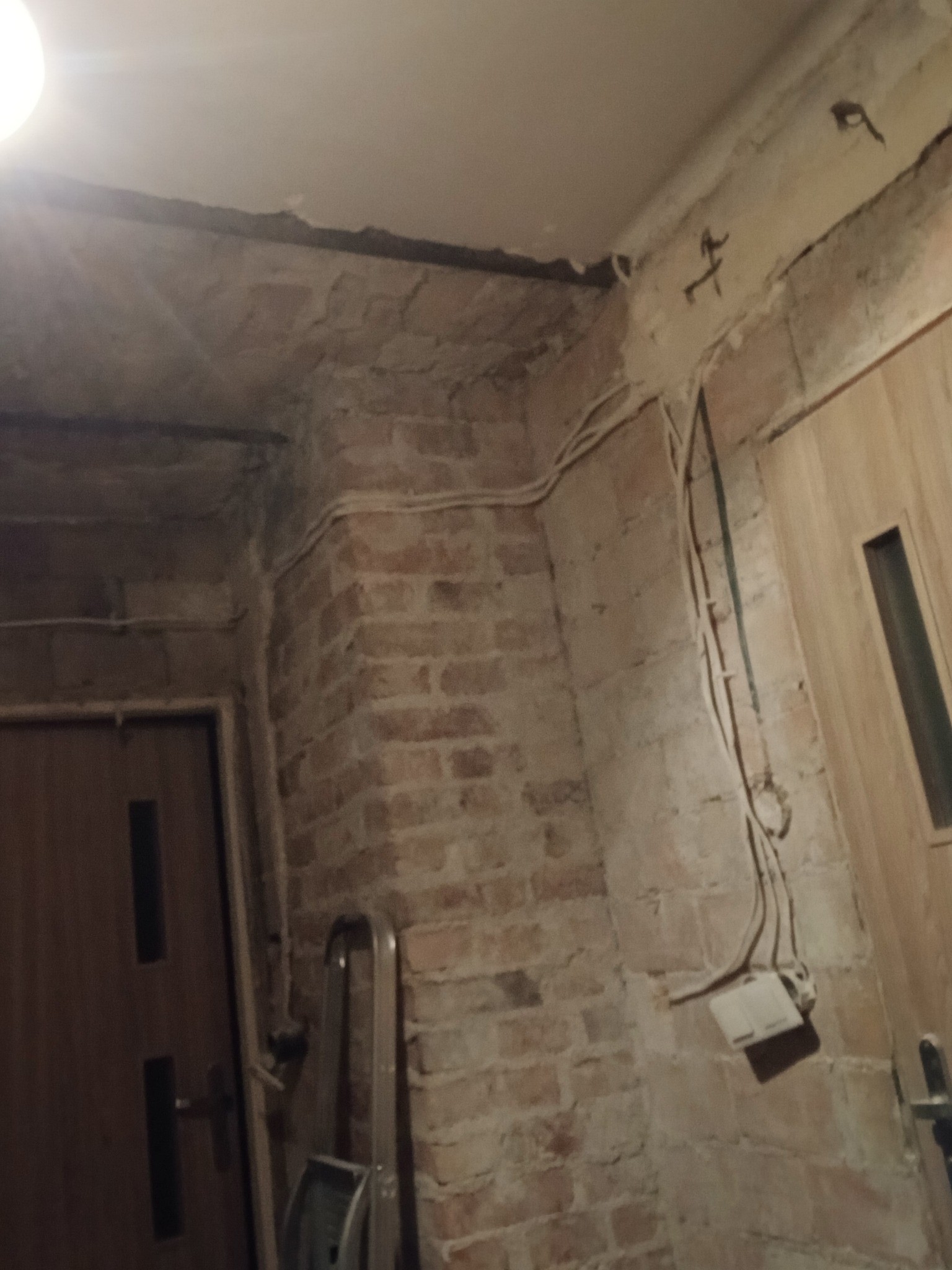 Ceglana ściana w trakcie remontu z widocznymi przewodami elektrycznymi, fragmentem drabiny i ościeżnicą drzwiową. Surowy stan wnętrza przed wykończeniem.