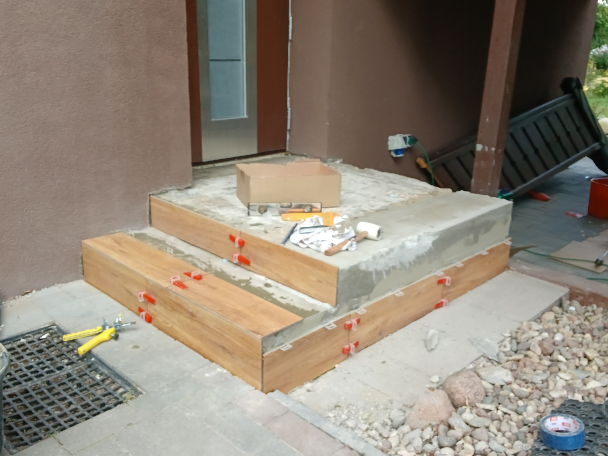 Montaż płytek imitujących drewno na schodach wejściowych, z użyciem klipsów poziomujących. Widoczne narzędzia glazurnicze i karton. Prace w toku, na zewnątrz budynku.