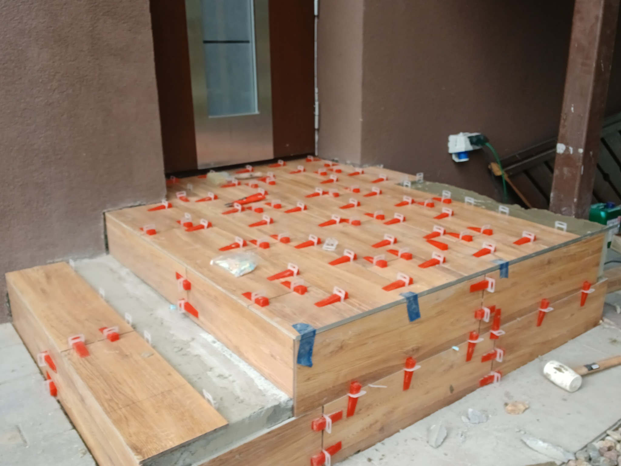 Układanie płytek imitujących drewno na schodach wejściowych, z użyciem czerwonych klipsów poziomujących. Widoczny młotek gumowy i paczka klipsów.