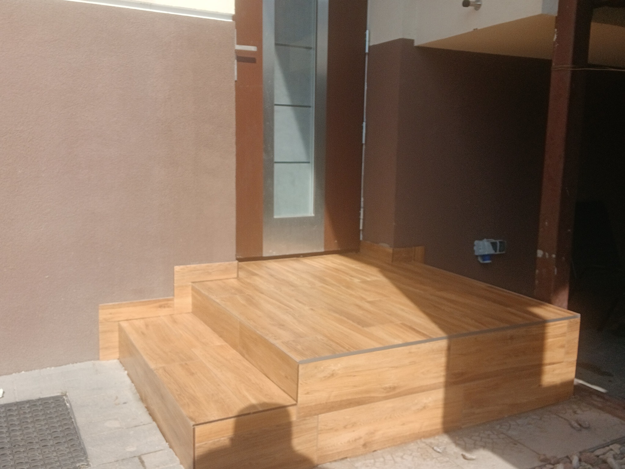 Wykończone schody wejściowe z płytek imitujących drewno, prowadzące do drzwi z szybą. Beżowa ściana i ciemnobrązowy filar w tle. Jasne, naturalne światło.