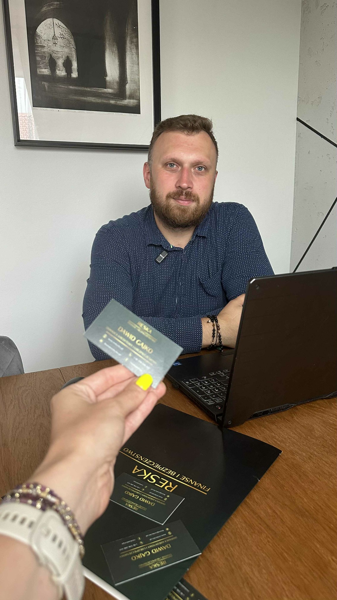 Doradca finansowy Dawid Gajko prezentuje wizytówkę. Na biurku laptop i materiały firmowe Reska Finanse i Bezpieczeństwo. W tle obraz w czarnej ramie.