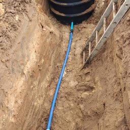 Przedsiębiorstwo Handlowo- Usługowe DREN-KAN. Ciapa Beata - Montaż szamba: czarny zbiornik z podłączoną niebieską rurą w wykopie, drabina oparta o brzeg. Widok z góry na instalację w ziemi.