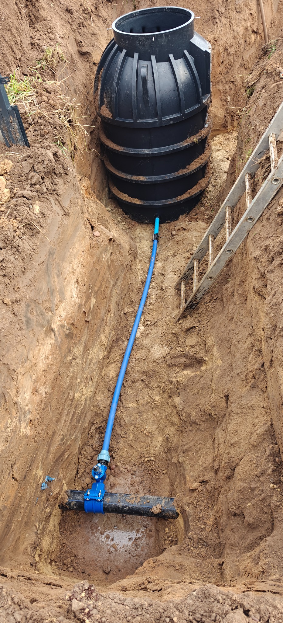 Montaż szamba: czarny zbiornik z podłączoną niebieską rurą w wykopie, drabina oparta o brzeg. Widok z góry na instalację w ziemi.
