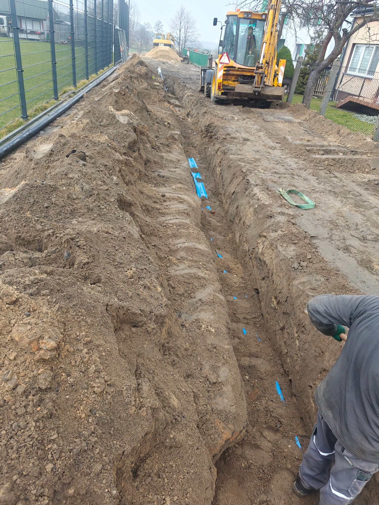 Wykop z ułożonymi rurami oznaczonymi niebieskimi taśmami, widoczny pracownik z łopatą i koparka w tle. Prace ziemne w trakcie realizacji.