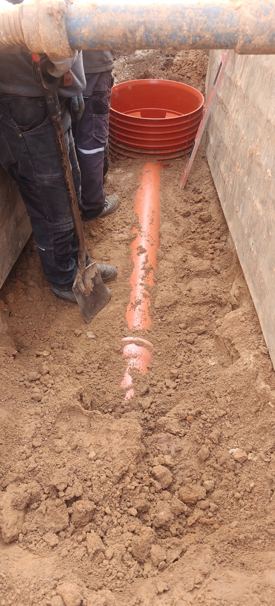 Pomarańczowa rura kanalizacyjna w wykopie, obok pracownik z łopatą i element studni kanalizacyjnej. Widok z góry na wykop.