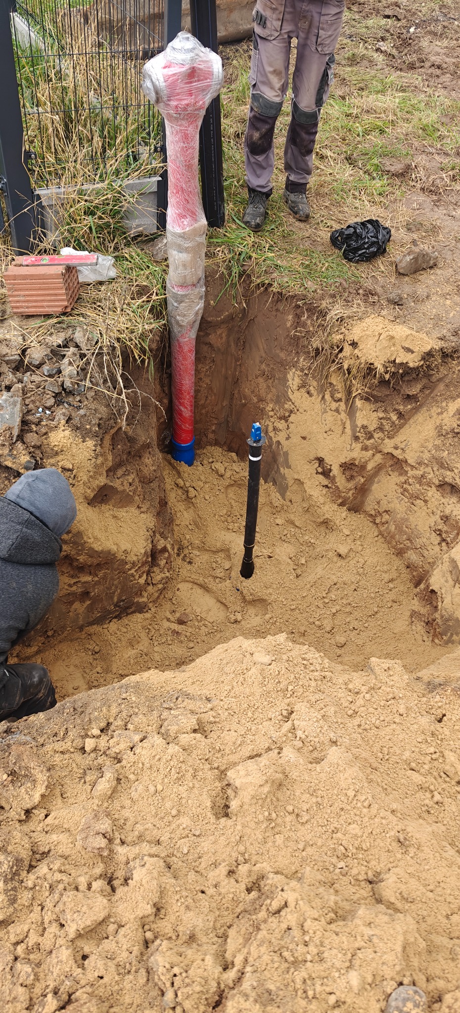 Czerwony hydrant owinięty folią w wykopie ziemnym, widoczne elementy instalacji wodnej i fragmenty odzieży robotników. Prace ziemne w toku, cegły obok wykopu.