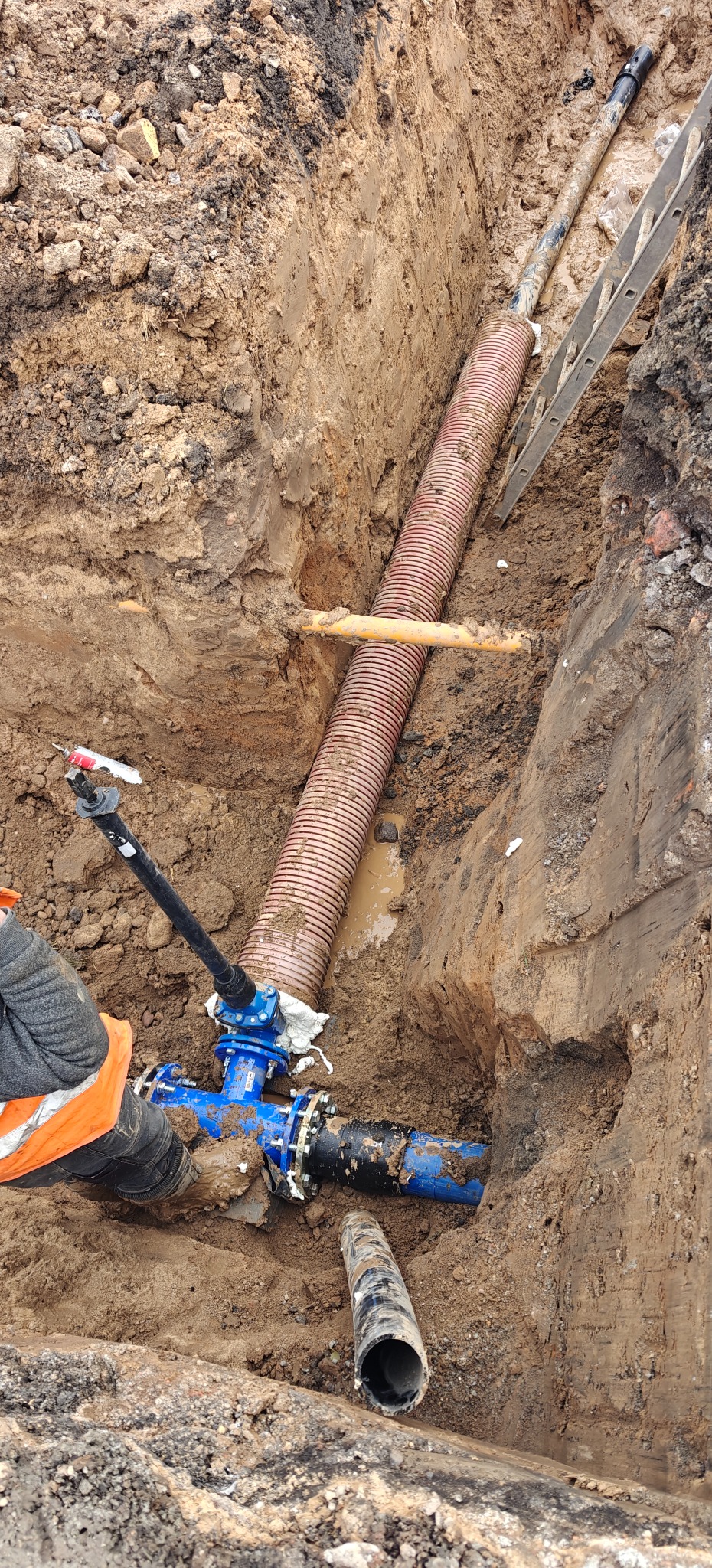 Wykonane przyłącze wod-kan w wykopie ziemnym z widocznymi rurami, złączkami i zaworem. Częściowo widoczna drabina i pracownik w odblaskowej kamizelce.