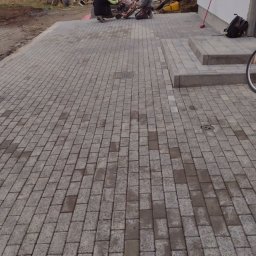 Blue Hare Szymon Zając - Dwóch pracowników docina kostkę brukową maszyną na nowo ułożonym podjeździe z szarej kostki, widoczne narzędzia, w tle budynek w trakcie wykończenia.