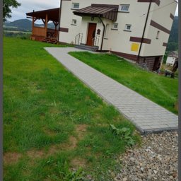 Blue Hare Szymon Zając - Ułożony chodnik z kostki brukowej prowadzący do domu z brązowymi akcentami, na zboczu wzgórza z widokiem na oddalone góry.