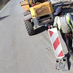 Blue Hare Szymon Zając - Żółta wywrotka na drodze, pracownik w odblaskowej kamizelce trzyma znak ostrzegawczy, w tle wykop i drzewa.