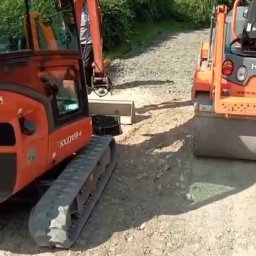 Blue Hare Szymon Zając - Pomarańczowy walec drogowy i koparka Kubota KX019-4 na żwirowej drodze, w tle zielona roślinność.