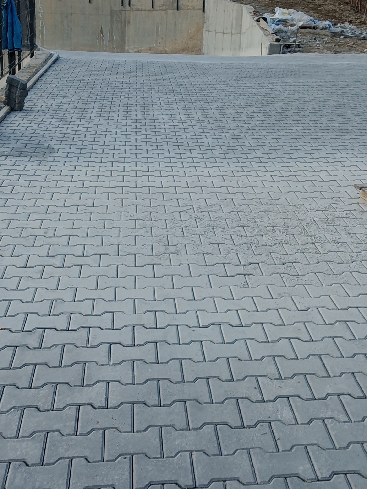 Ułożona szara kostka brukowa w wzór jodełki na podjeździe, z widocznymi pozostałościami piasku na powierzchni, z betonowym murem w tle i stosem kostki po lewej stronie.