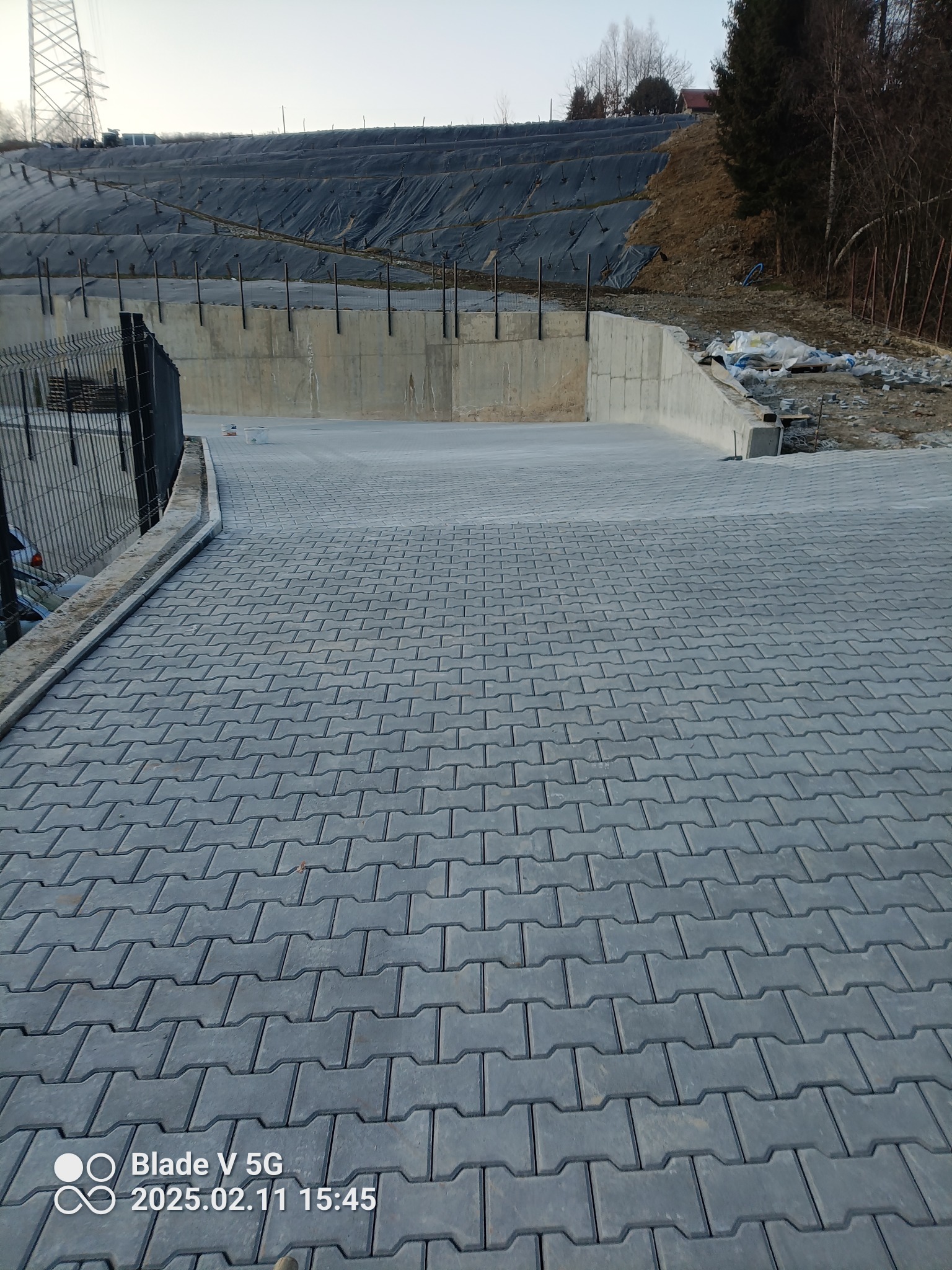 Ułożona szara kostka brukowa na utwardzonym placu z betonowym murem oporowym i zabezpieczonym skarpą w tle.